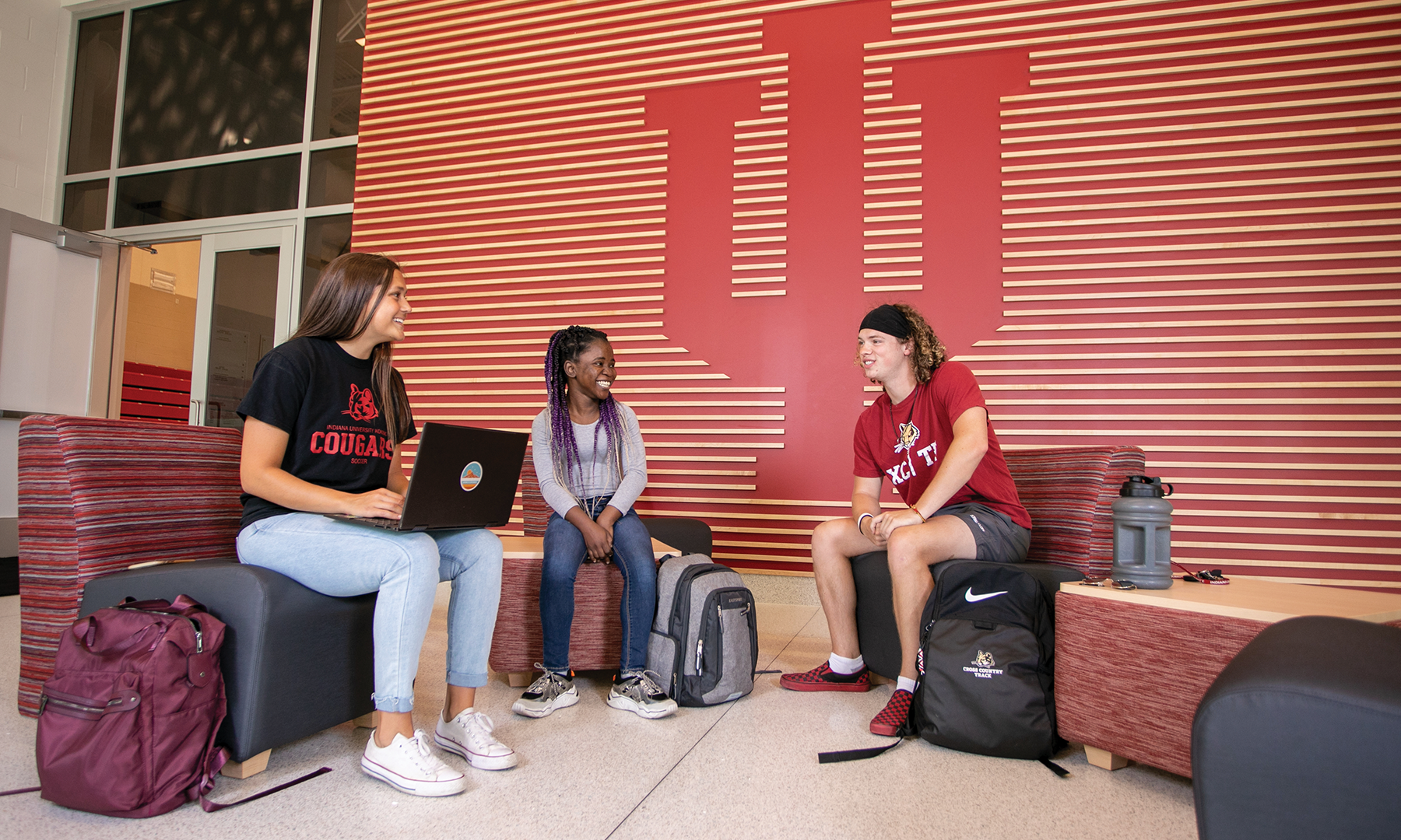 two female and one male student sitting in front of an IU trident wall in the Student Activities and Events Center lobby