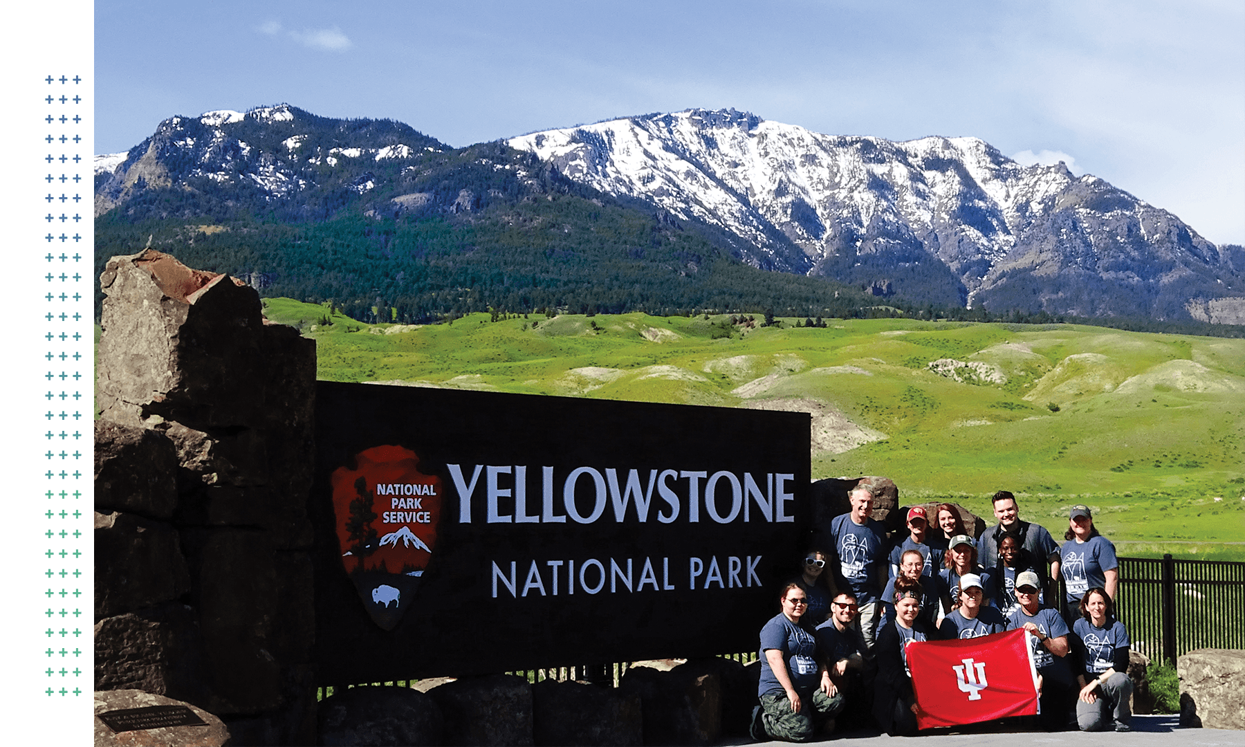 group of students posing with an IU flag next to a sign that reads Yellowstone National Park with snowcapped mountains in the background