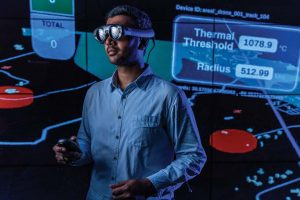 A man wearing VR goggles stands in front of a large monitor displaying data visualizations.