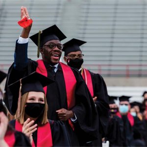 Students wearing their graduation caps and gowns, as one student stands and cheers with his hand raised.