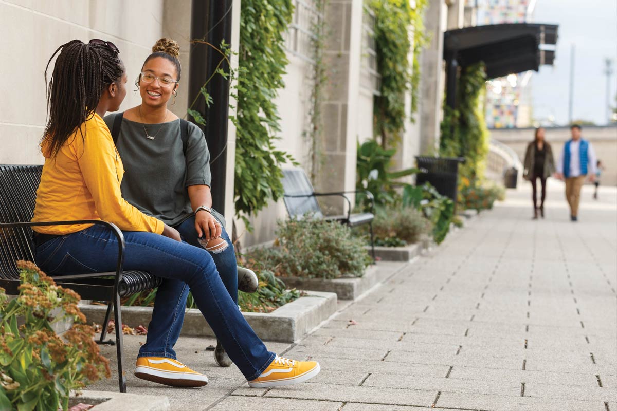 Two femail students sit on a bench on the downtown Indy canal with vines amid vines and grasses