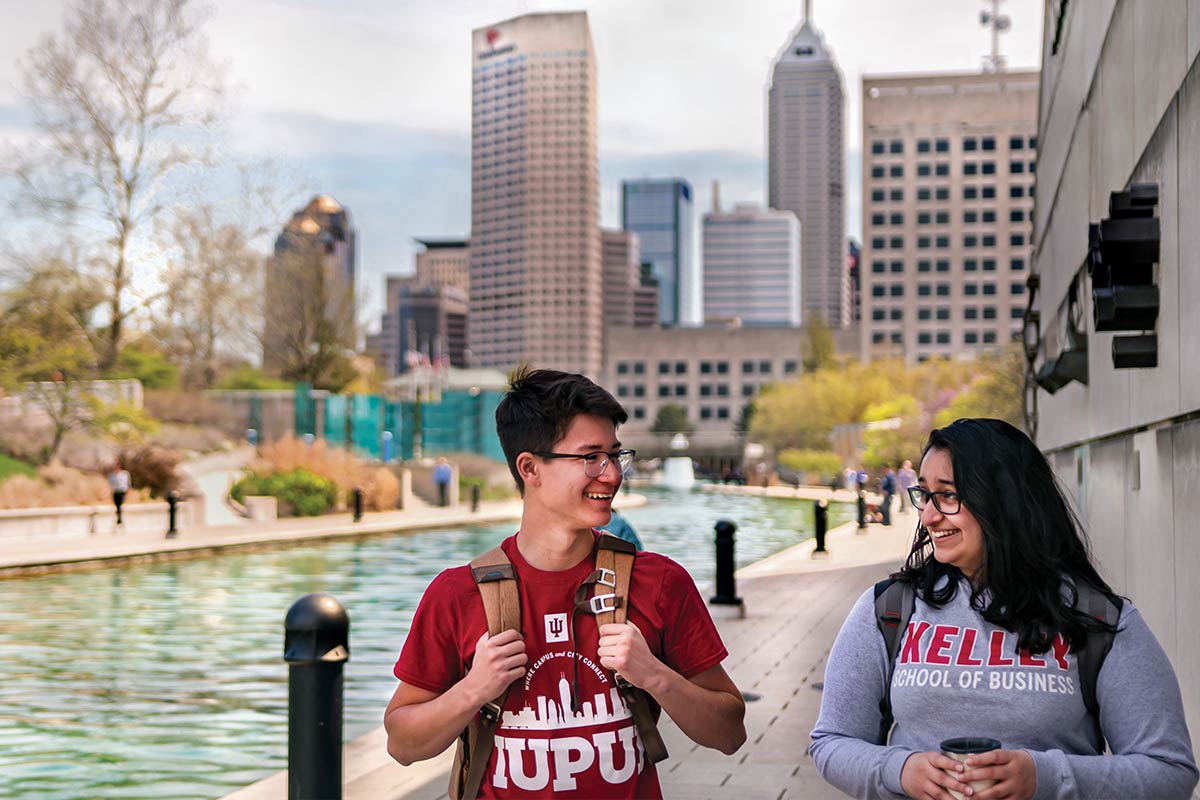 Two students with backpacks walk along the downtown canal with the skyline in view behind them.