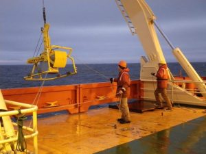Photo showing Box corer that two crew members are lowering from the ship to the sea floor.