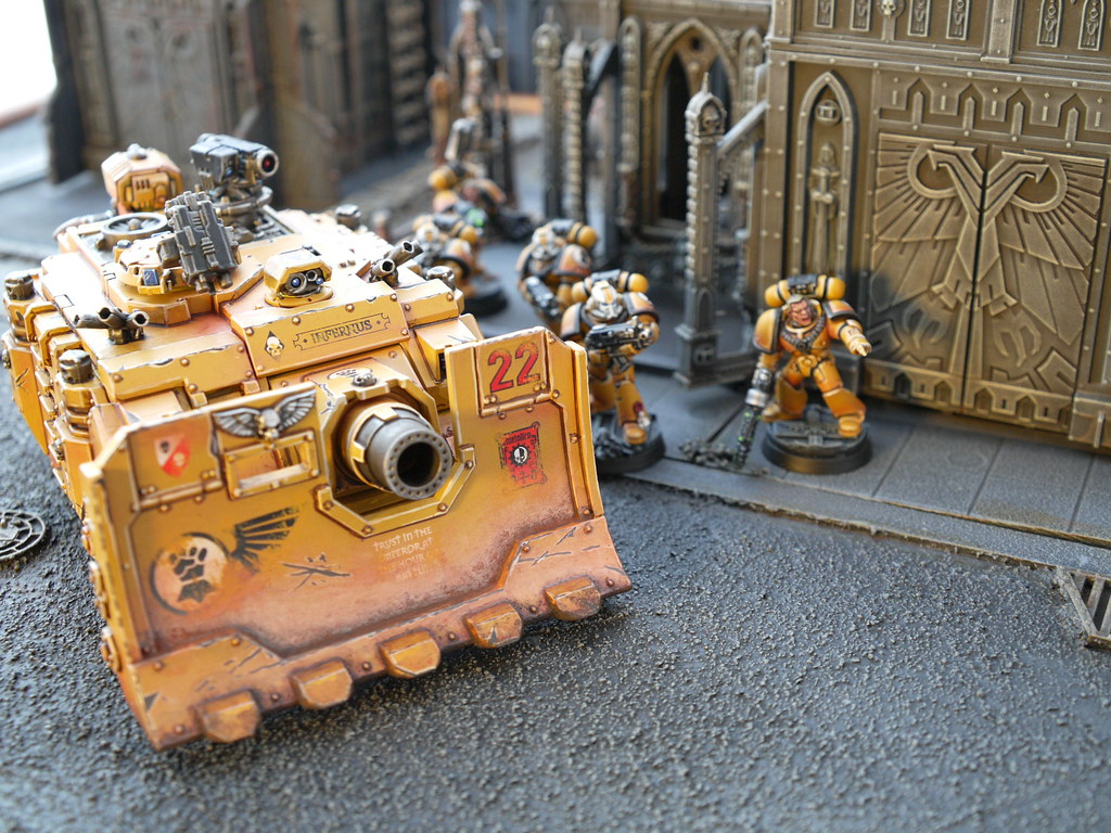 Image of Iron Fist miniatures. Yellow tank with three small men in yellow armor behind it. A large carved building wall is behind the miniatures.