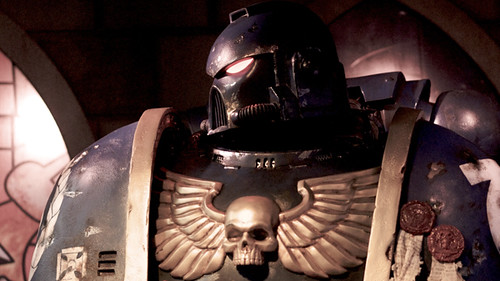 Close up image of a Space Marine. Man in blue power armor with a gold skull with wings on his chest.