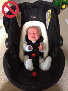 Infant secured in a car seat with additional support cushion, highlighted by a red circle with a line through it, indicating that the use of extra support is not recommended or prohibited