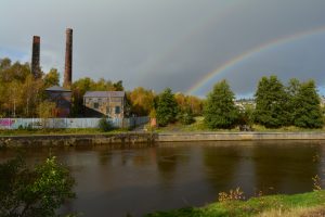 View of a building and smokestack from across a river with a rainbow in the sky.