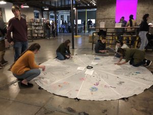 People are kneeling on a round, white parachute on the floor of an art gallery and writing or drawing their responses to the prompt, "what does environmental justice mean to you?"