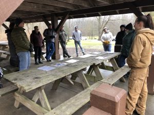 A group of people are gathered around a wooden picnic table in a covered pavilion with trees and grass visible in the background and there are laminated pages spread out on the table.