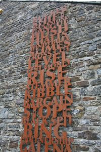 A rusted steel metal sculpture of cut out letters forming words and mounted on a stone wall.