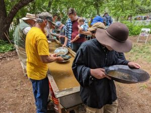 visitors holding round pans with water and sediment stand around a wooden trough set up to allow visitors to experience gold mining.