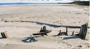 photograph of a sandy beach with remnants of a rusted vehicle sticking up through the sand and the ocean shoreline is visible in the background.