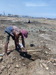 A man working with a pickax is digging through a barren area of dirt and e-waste materials.