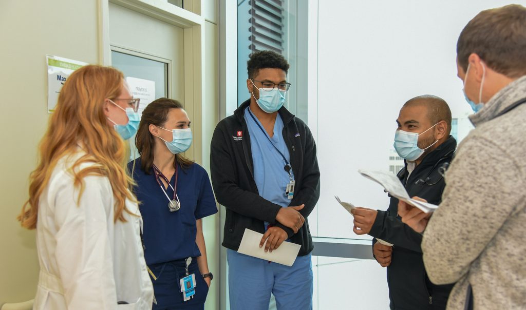 A diverse group of young physicians meet in a hospital hallway. Each is wearing a blue surgical mask.