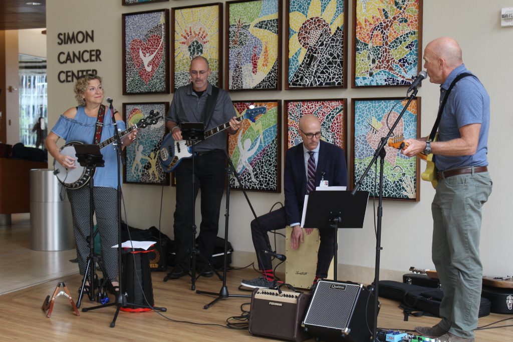 Four people playing various instruments, including guitars, a banjo, and a drum, perform music in front of a colorful background.