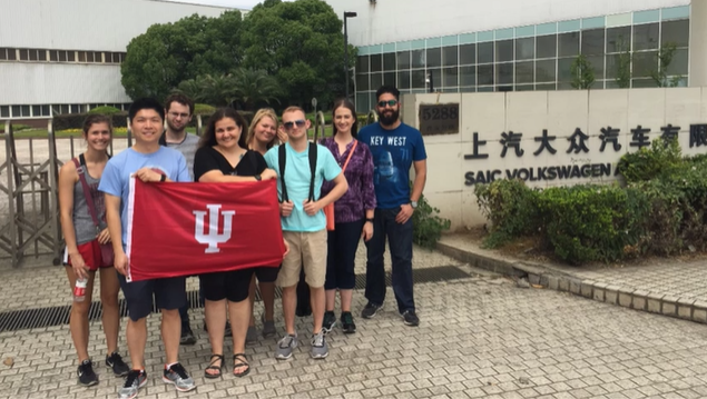 A photo of a group of people posing with a "Indiana University" flag in front of a building.