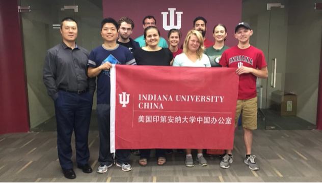 A photo of a group of people posing with a "Indiana University" flag in front of a wall with the Indiana University logo on it.