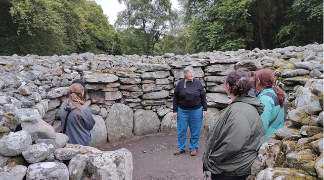 A photo of six people standing in ancient Scottish burial ruins in conversation.