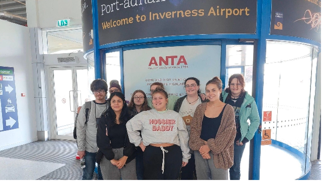 A photo of nine students in front of a "Welcome to Inverness Airport" sign in the airport.