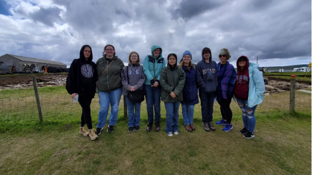 A one photo of nine people posing in front of an archaeology site.