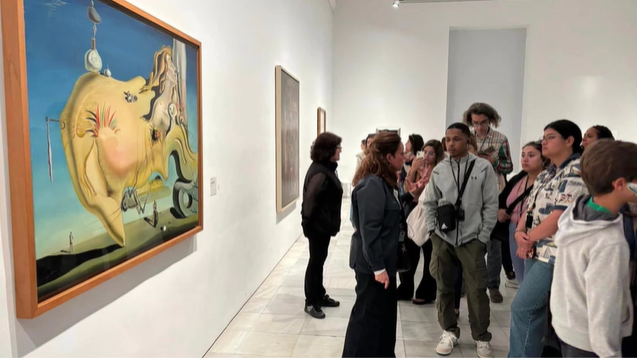 A photo of a group of people listening to a person speak in front of a painting.