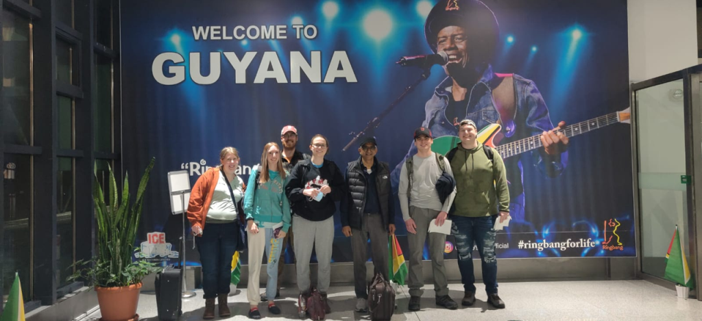 Image of a group of people posing in front of a wall that reads "Welcome to Guyana".