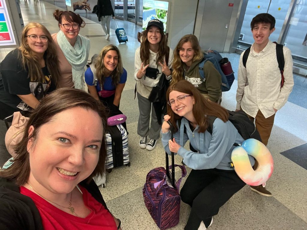 Image of eight people posing for a selfie with luggage in an airport.