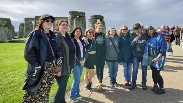 A photo of nine people posing in front of Stonehenge.