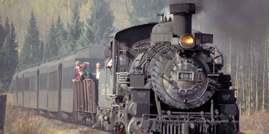 A steam engine and several passenger cars are shown traveling down a train track. The train has some people on board.
