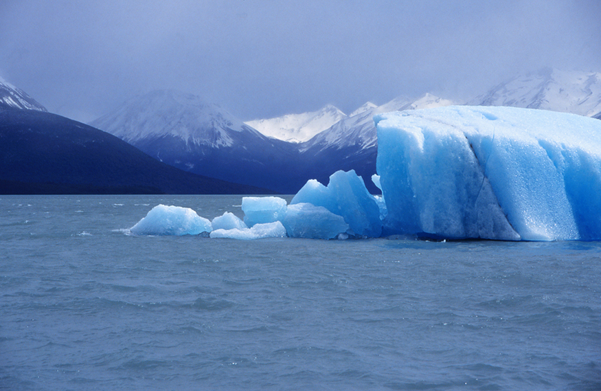 The figure shows some blue-colored icebergs floating in the water beneath snow-capped mountains and a cloudy sky. Some of the icebergs at front are melting.