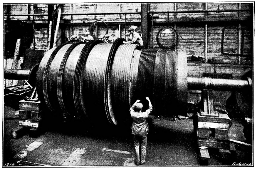 An old photo of a steam turbine at a turbine production plant. People are shown working on the turbine.