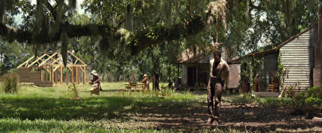 Delineating the plantation household, the panoramic shot shows Solomon as the hanged man with the slave cabins in the background and the slaves going about their daily routines (screen capture by author).