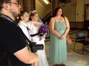 Two women standing next together, one woman dressed in a white wedding gown holding purple flowers, another is wearing a white tuxedo.  Male photographer and a woman in a green dress stand on either side of the couple.