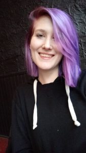 Caucasian woman smiling with lavender-colored, shoulder-length hair and wearing a black sweater.