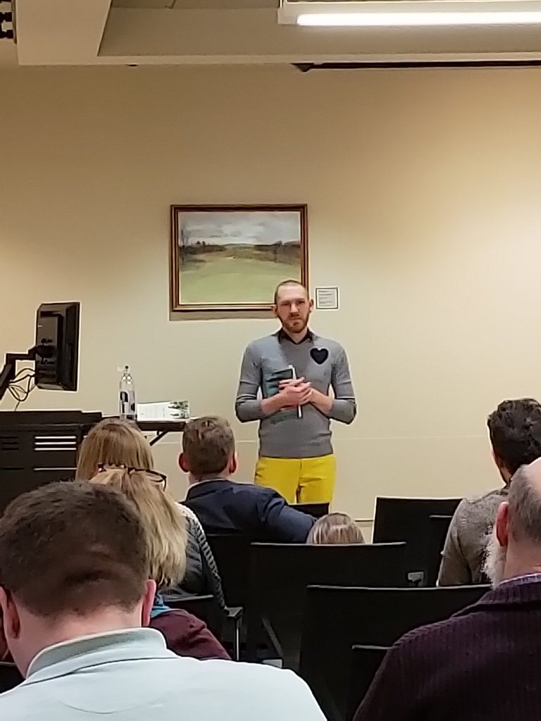 Image of of Stephan Mills at his poetry reading. Mills is standing at the front of the room, holding a book, wearing a gray sweater with a heart shape patch on his left shoulder and yellow pants.