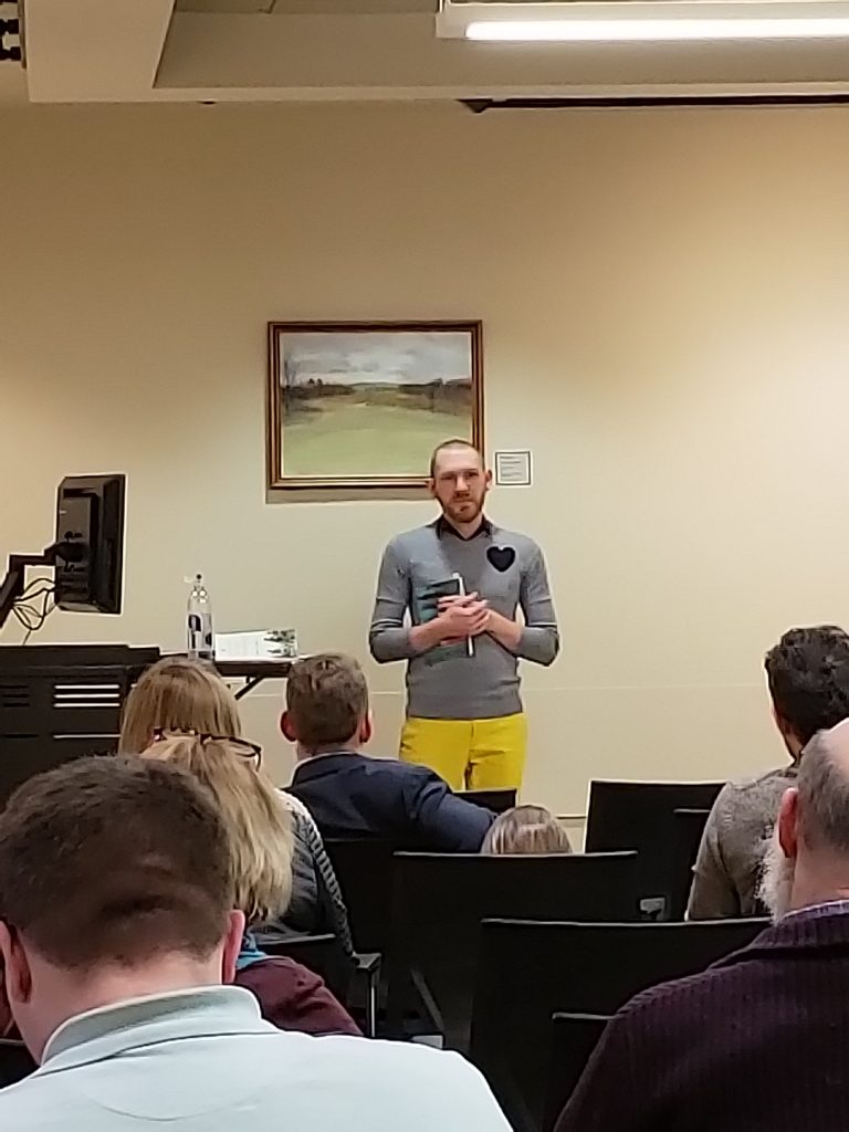 White caucasion male standing at the front of the room, holding a book, wearing a gray sweater with a heart shape patch on his left shoulder and yellow pants.