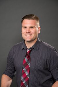 Work photo of Josh Tolbert. Caucasian male wearing gray button down shirt and red and gray plaid tie.