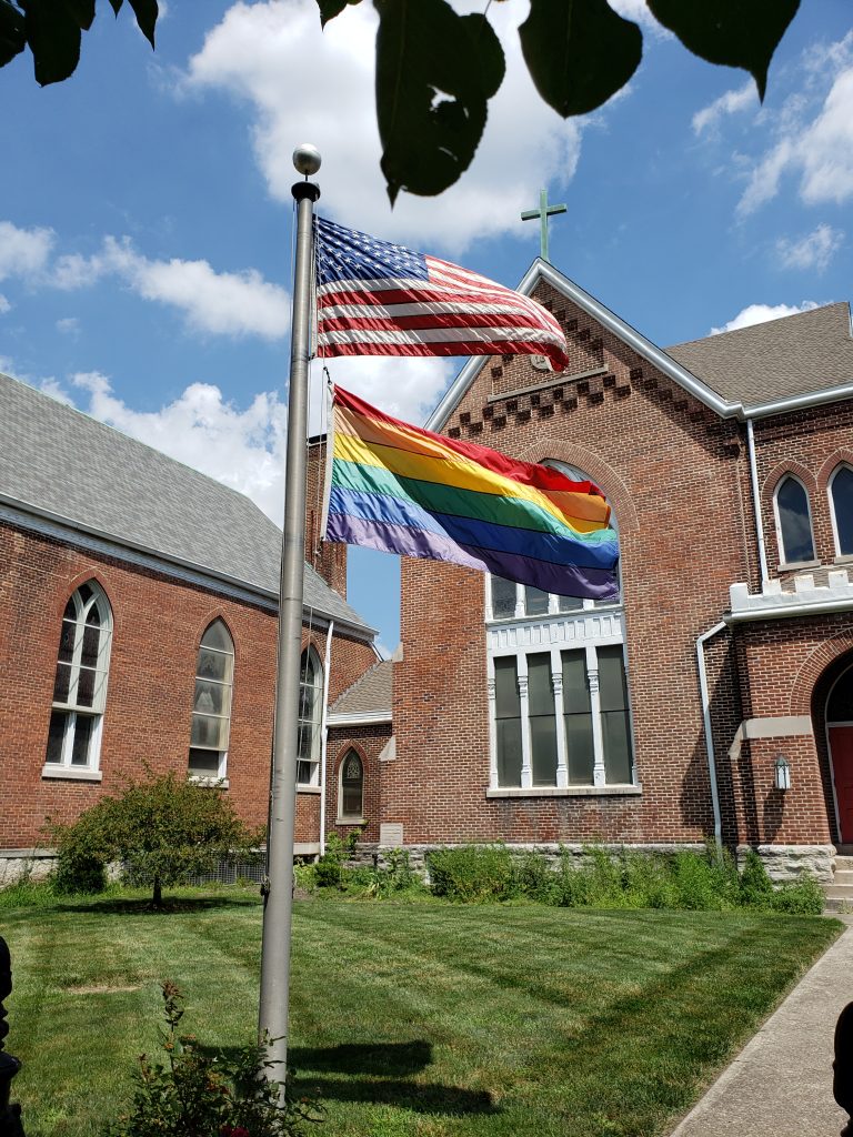 Picture of St. Paul's Episcopal Church. The image has the American Flag and a rainbow flag flying together in front of the church.