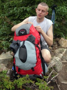 A hiker kneeling over a large backpack in a wooded, rocky outdoor area.