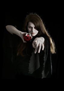 A young person with long hair wearing dark clothing, holding a red object, and standing against a black background.