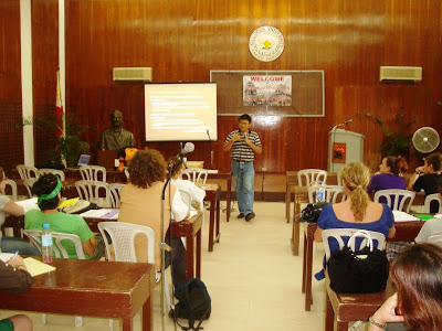 A room with people sitting at desks while a person stands in front speaking into a microphone.