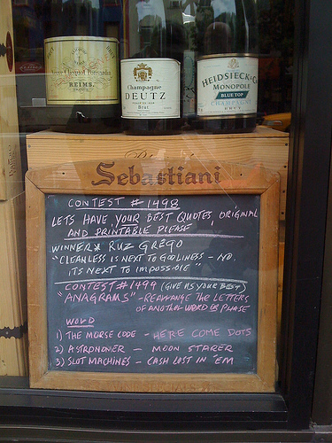 A window display of wine bottles placed on top of a box. A chalkboard containing handwritten information about a store contest is propped up in front of the box.