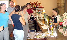 A group of people standing in front of a counter containing wine bottles and glasses for sampling. A salesperson behind the counter talks to some of the people.
